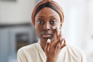 Dark skinned women putting cream on her cheek with her finger while looking in the mirror